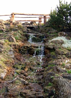 Ein kleiner Bergbach fließt einen Hang hinab auf der Kuppe befindet sich eine rustikales Holzgitter über grünen Bergpflanzen
